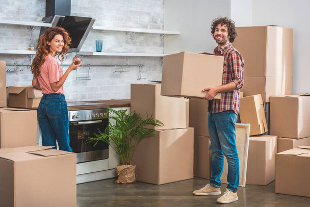 smiling couple unpacking cardboard boxes at new kitchen and looking at camera - Photo, Image