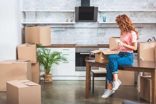 side view of woman holding cup of coffee and unpacking cardboard box on table at new home - Photo, Image