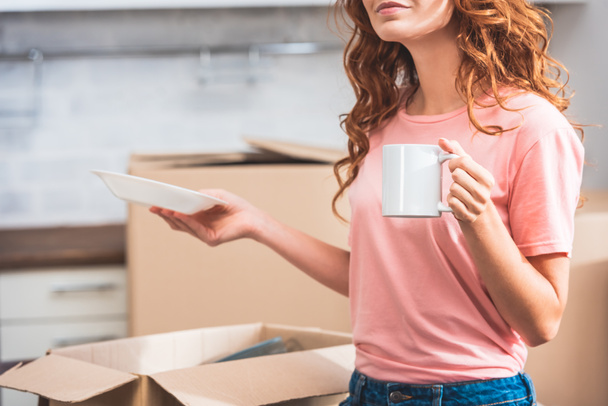 обрізане зображення жінки з кучерявим рудим волоссям, що тримає чашку кави і тарілки в новому будинку
 - Фото, зображення