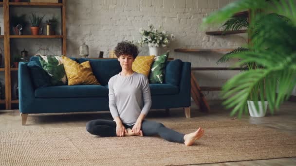 Flexible girl is bending forward in seated position and twisting her body enjoying yoga and activity sitting on floor in beautiful apartment. People and health concept. - Кадри, відео