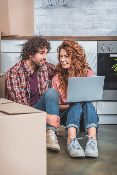 couple with curly hair sitting with laptop near cardboard boxes on floor in new kitchen - Photo, image