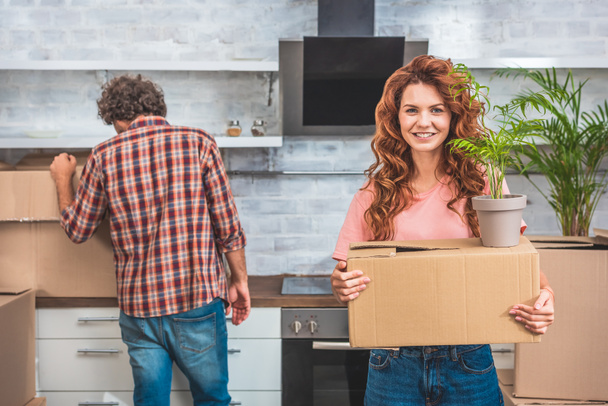 couple unpacking cardboard boxes at new home, girlfriend holding box with plant and looking at camera - Photo, Image