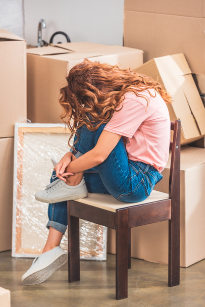 woman with curly red hair sitting on chair near cardboard boxes at new home - Фото, изображение