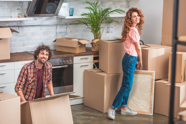 smiling couple unpacking cardboard boxes at new home and looking at camera - Photo, Image