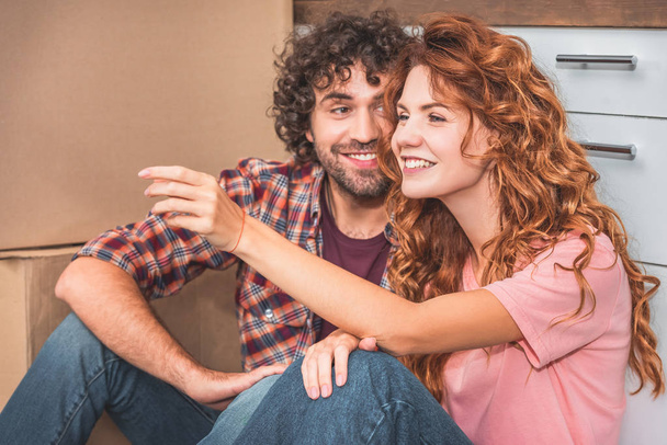 cheerful couple sitting on floor near cardboard boxes in new kitchen, girlfriend gesturing and looking away - Photo, Image