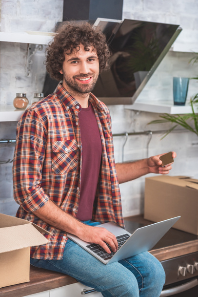 smiling handsome man with curly hair sitting with laptop and credit card on kitchen counter at new home - Foto, Imagen