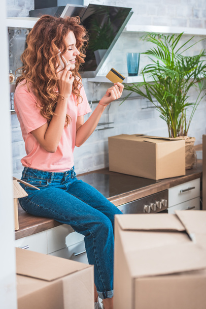 счастливая молодая женщина, держащая кредитную карту и разговаривающая по смартфону во время переезда в новую квартиру
 - Фото, изображение