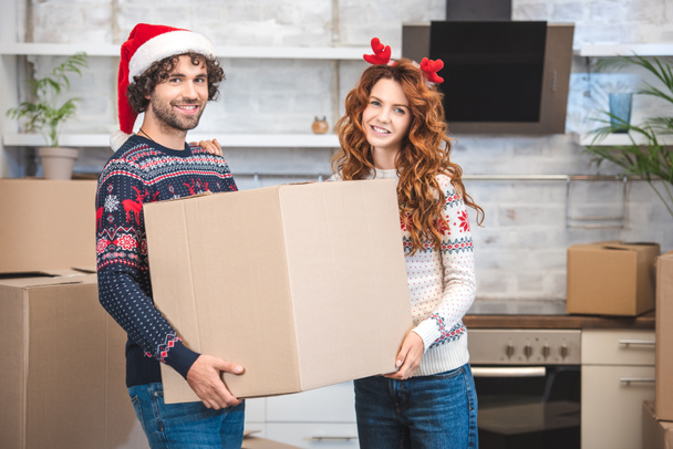 happy young couple holding cardboard box and smiling at camera while relocating in new apartment at christmastime  - Foto, Bild