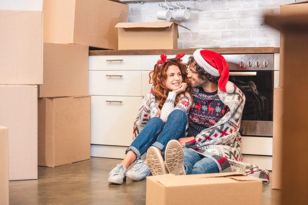 glückliches junges Paar lächelt einander zu, während es zur Weihnachtszeit zwischen Kartons sitzt - Foto, Bild