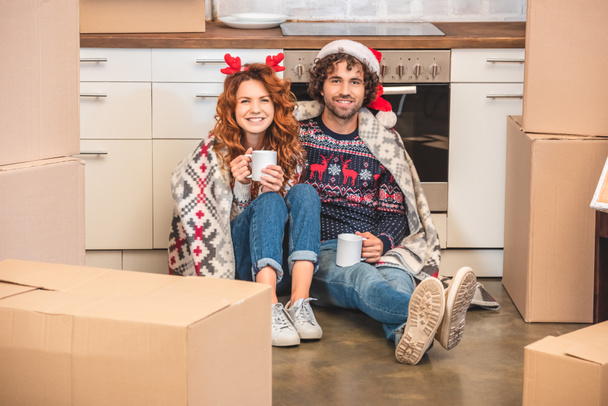 heureux jeune couple tenant des tasses et souriant à la caméra tout en étant assis entre les boîtes de carton à Noël
 - Photo, image