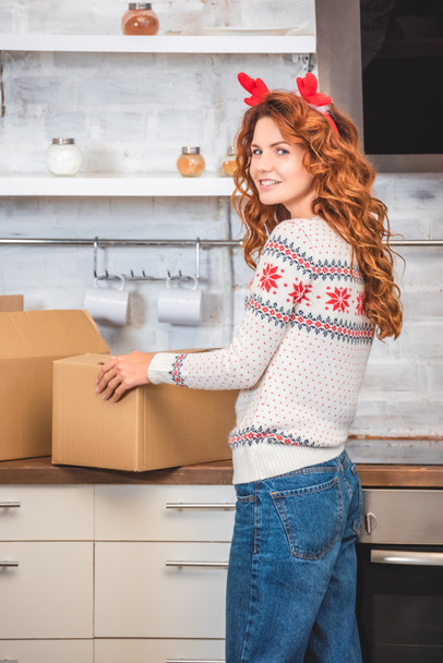 beautiful young woman in antlers headband holding cardboard box and smiling at camera while relocating in new apartment  - Foto, Imagem