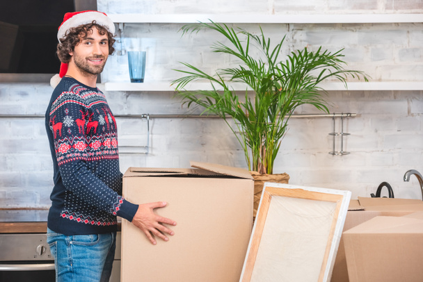 handsome happy young man in santa hat holding cardboard box and smiling at camera during relocation - Photo, Image