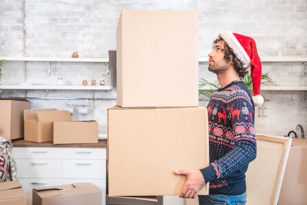 side view of young man in santa hat holding cardboard boxes during relocation - Foto, Imagem