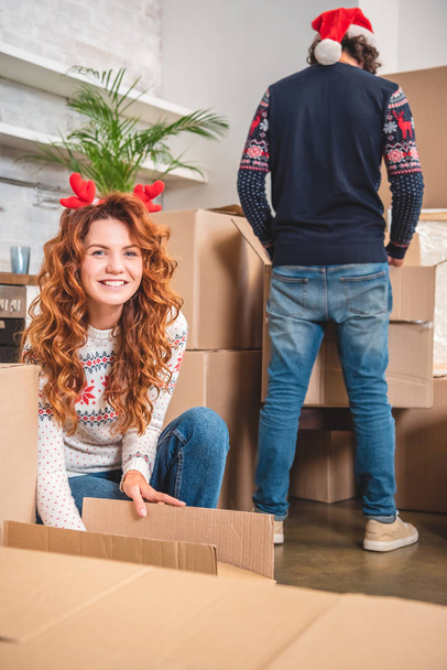 glückliche junge Frau im Geweih Stirnband auspackt Kartons und lächelt in die Kamera, während Mann mit Weihnachtsmütze hinter steht - Foto, Bild
