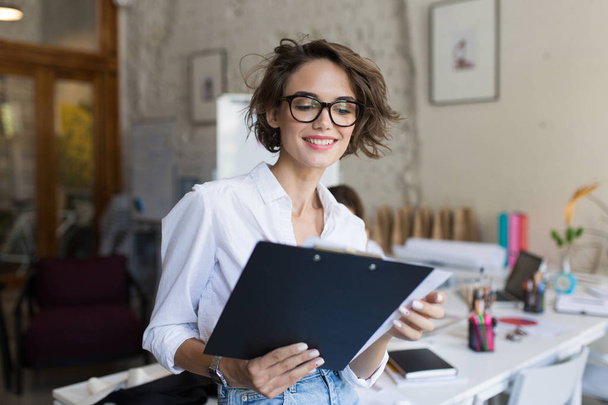 Young beautiful smiling girl with short curly hair in eyeglasses and white shirt holding folder in hands at work in modern cozy office - 写真・画像