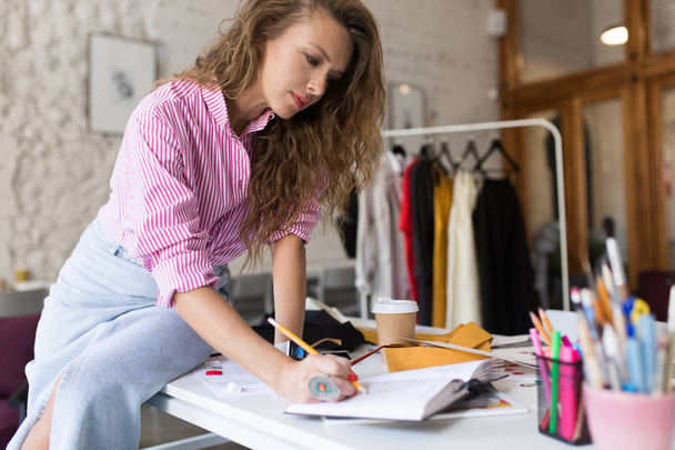 Νεαρή όμορφη γυναίκα στο ριγέ πουκάμισο και τζιν φούστα με μεγάλο δαχτυλίδι στο δάχτυλο γράφει σημειώσεις στο σημειωματάριο στην εργασία στο μοντέρνο άνετο γραφείο - Φωτογραφία, εικόνα