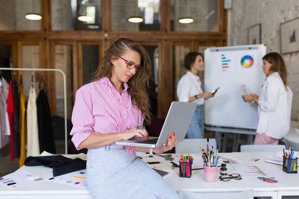 Stijlvolle jongedame in gestreepte shirt en denim rok en brillen leunend op Bureau dromerig werken op de laptop aan het werk in moderne gezellig kantoor - Foto, afbeelding