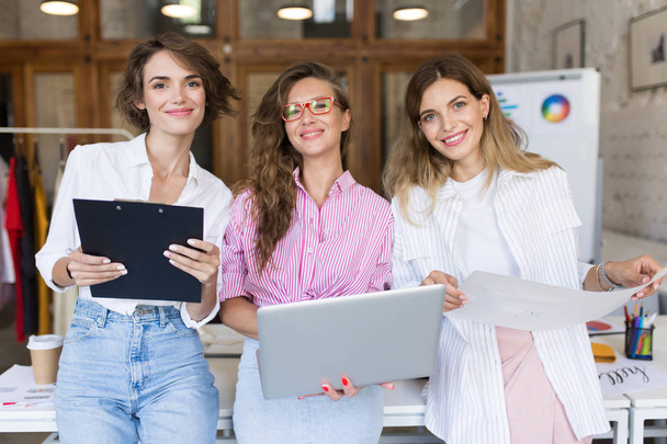Group of young beautiful stylish women with laptop and folder happily looking in camera working together in modern cozy office - Photo, Image