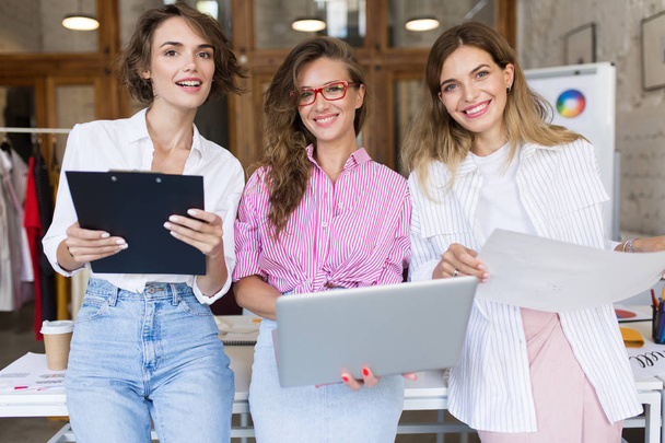 Groep van jonge mooie stijlvolle vrouwen met laptop en een map vol vreugde op zoek in camera werken samen in moderne gezellig kantoor. Mooie meisjes besteden tijd op het werk - Foto, afbeelding