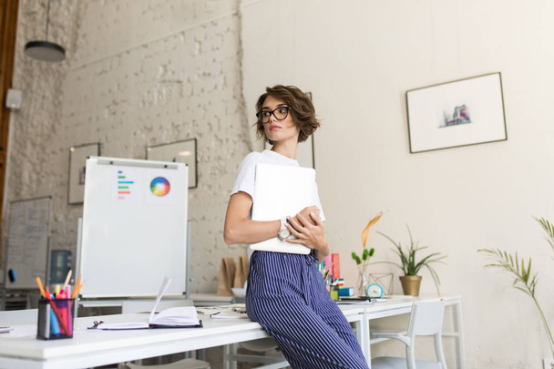 Красивая девушка с короткими вьющимися волосами в очках и белой футболке мечтательно смотрит в сторону с ноутбуком в руках, опираясь на стол на работе в современном офисе
 - Фото, изображение