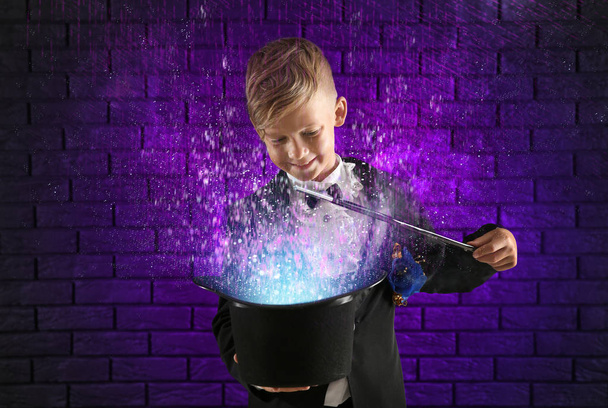 Cute little magician showing trick with hat against dark brick wall - Photo, Image
