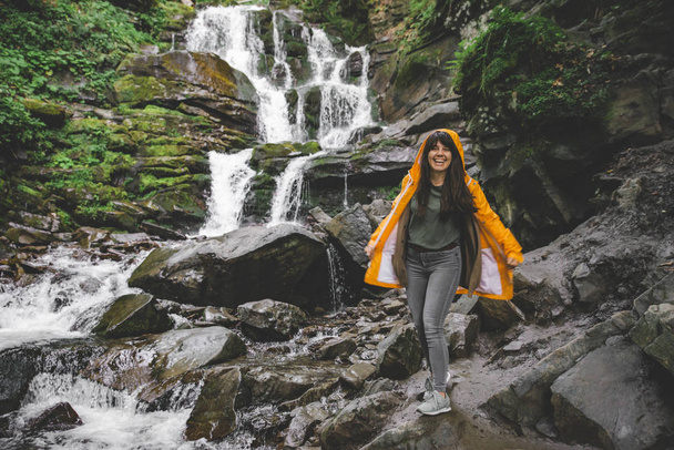 mulher de pé em capa de chuva amarela e olhando para cachoeira. conceito de caminhadas
 - Foto, Imagem