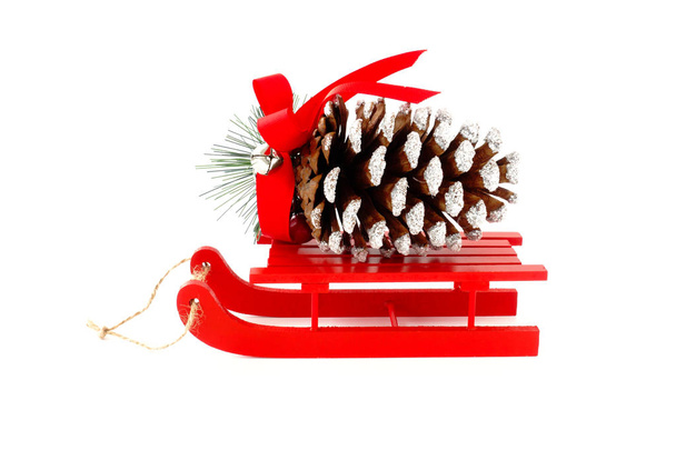 Olbrzymie Boże Narodzenie pinecone ozdobione srebrny brokat, bell i czerwoną wstążką na czerwone drewniane sanki na białym tle. - Zdjęcie, obraz