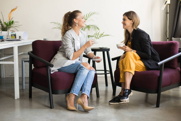 Two beautiful stylish women happily talking holding cups of coffee in hands spending time together in modern comfortable office - Photo, Image