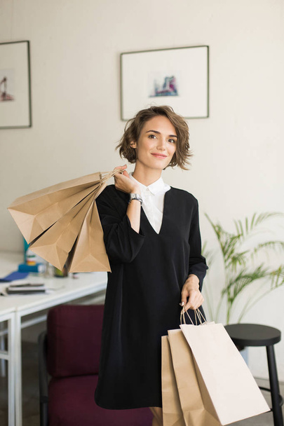 Beautiful smiling girl in black dress dreamily looking in camera holding paper packets in hands in modern office - Foto, afbeelding
