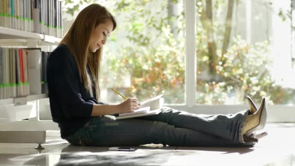 θηλυκό φοιτητής κάθεται στο πάτωμα, στη βιβλιοθήκη, ανάγνωση, βιβλίο και σημειώσεις - Πλάνα, βίντεο