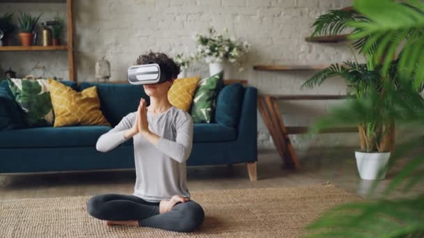 Slender girl in virtual reality glasses is meditating sitting on floor at home in lotus position and enjoying personal practice. Modern technology and yoga concept. - Materiał filmowy, wideo