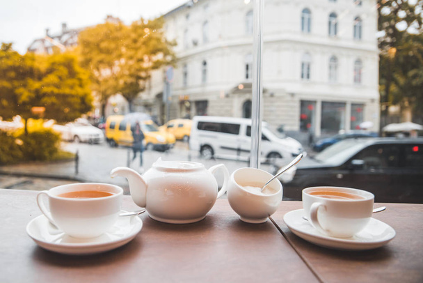 kaksi kuppia teetä ja valkoinen vedenkeitin kahvilapöydässä iso lasi-ikkuna. syyskausi
 - Valokuva, kuva