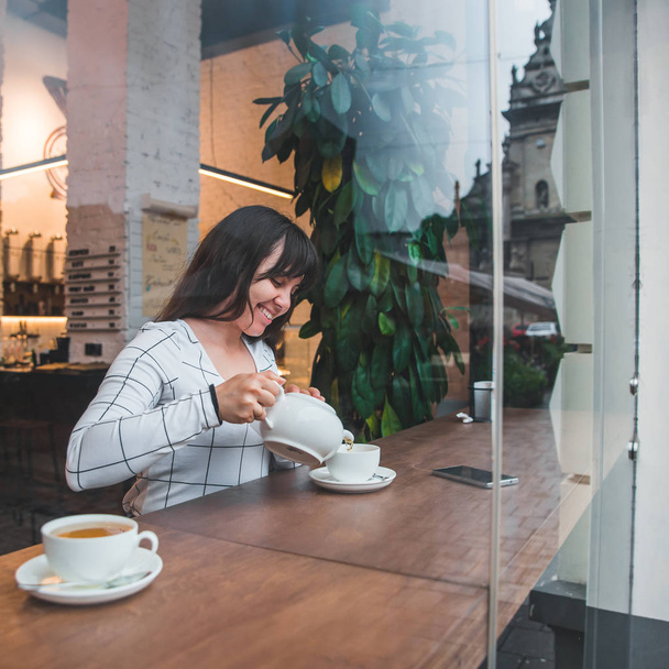 Frau gießt Tee in die Tasse im Café. Blick durch Glasfenster - Foto, Bild