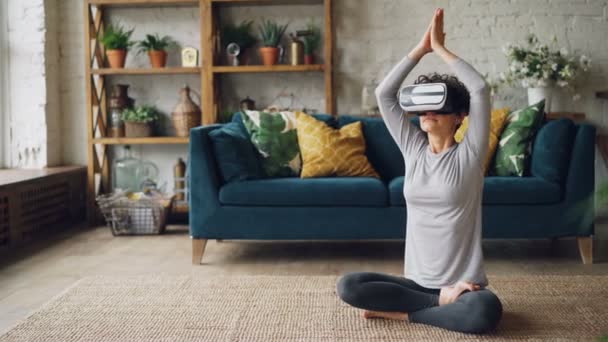 Slim young lady in artificial reality glasses is meditating at home in lotus pose with hands in namaste enjoying simulation and relaxing. Modern technology and experience concept. - Filmagem, Vídeo