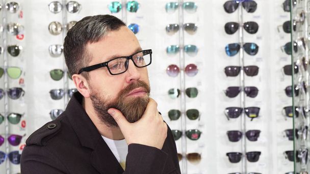 Homem maduro barbudo esfregando sua barba cuidadosamente escolhendo novos óculos
 - Foto, Imagem