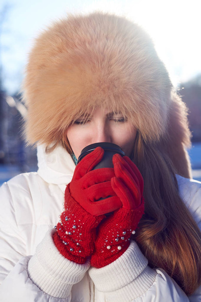 Pelirroja chica flaca, vestida con sombrero de piel de zorro y chaqueta blanca, bebiendo café para ir en invierno día frío
.  - Foto, imagen