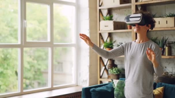 Happy young lady is wearing trendy artificial reality glasses standing at home in loft style apartment and gesturing. Curly-haired girl is wearing casual clothing. - Filmagem, Vídeo