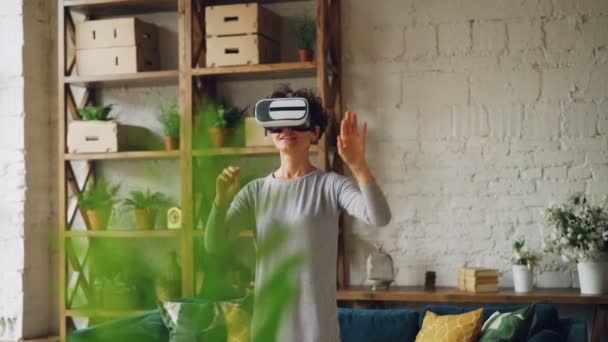 Slender girl is using virtual reality glasses at home standing in loft style room and moving hands playing game. Modern technology, leisure and new experience concept. - Séquence, vidéo