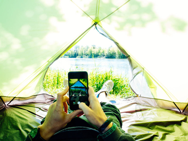 Photo of a man laying in tourist tent with a view of the forest and lake in sunny summer day. - Foto, imagen