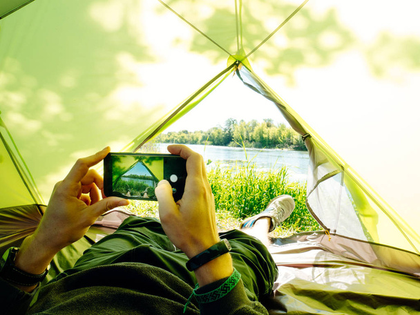 Photo of a man laying in tourist tent with a view of the forest and lake in sunny summer day. - Foto, imagen