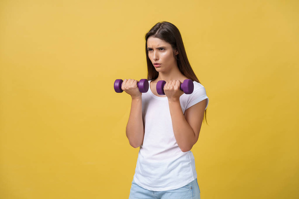 Feeling exhausted. Frustrated young woman in white shirt exercising with dumbbells and serious look while standing isolated on yellow background - Photo, Image