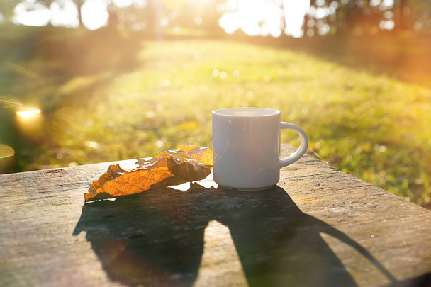 М'який фокус білого кавового горнятка і сухого листя на дерев'яному з сонячним світлом і світловим відблиском вранці
. - Фото, зображення