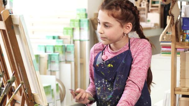 Красивая маленькая девочка рисует на мольберте масляными красками в художественной студии
 - Фото, изображение