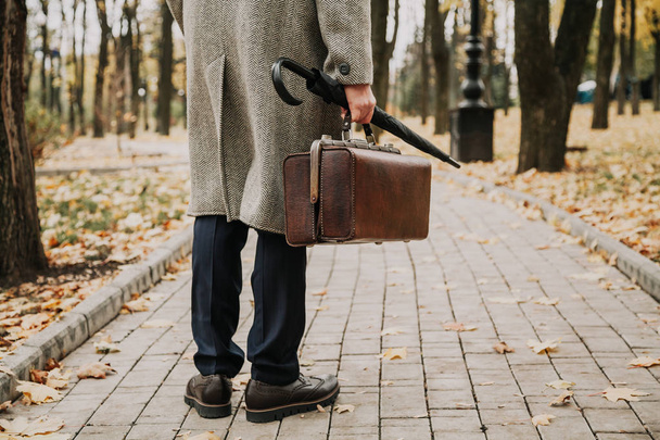 Caballero espera. Un hombre con zapatos elegantes y un abrigo blanco está de pie en una estrecha avenida del parque de otoño. Un paraguas y un ba
 - Foto, imagen