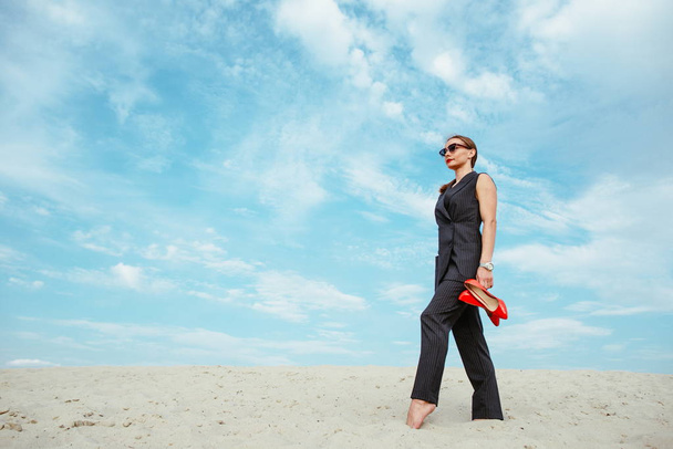 full height portrait of stylish white young woman in suit, sunglasses, red lipstick standing with red glossy high heels shoes in her hands outdoor in desert - Fotó, kép