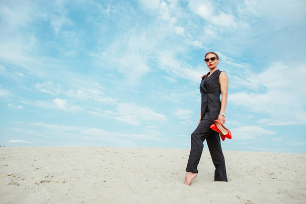 volledige hoogte portret van stijlvolle witte jonge vrouw in pak, zonnebril, rode lippenstift permanent met rode glanzende hoge hakken schoenen in haar handen buiten in woestijn - Foto, afbeelding
