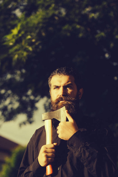 handsome bearded young man hipster with long beard and mustache has stylish hair on serious face sunny day outdoor holding sharp axe or ax on green natural background. - Valokuva, kuva