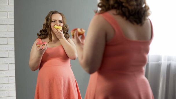 Unhappy fat female eating donuts in front of mirror, nutrition disorder issue - 写真・画像