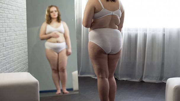 Overweight girl checking skin in front of mirror, looking at cellulite on body - Foto, afbeelding