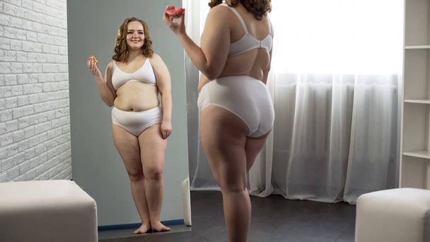 Overweight girl admiring mirror reflection, eating sweet donut, body positive - Valokuva, kuva
